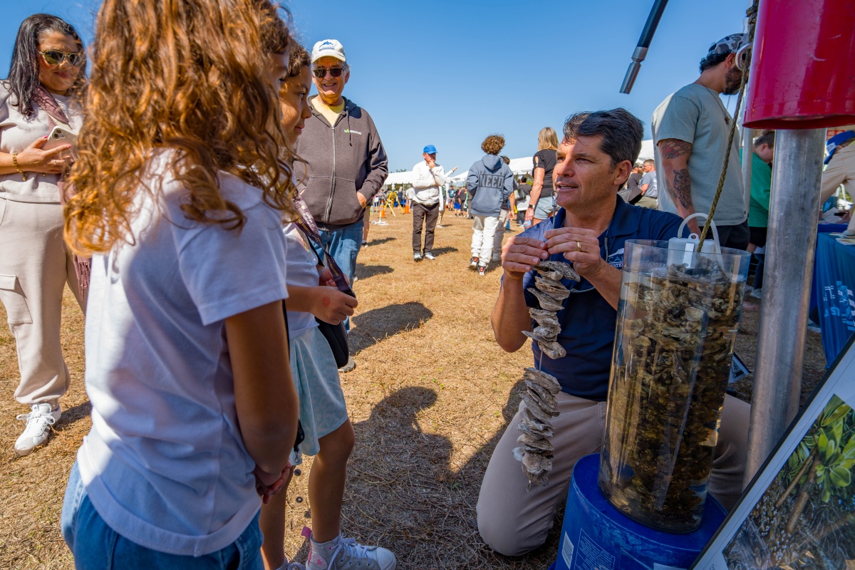 A man kneels beside a tube of oyster shells and holds up a string of oysters to show two young girls at the Waterways Festival.